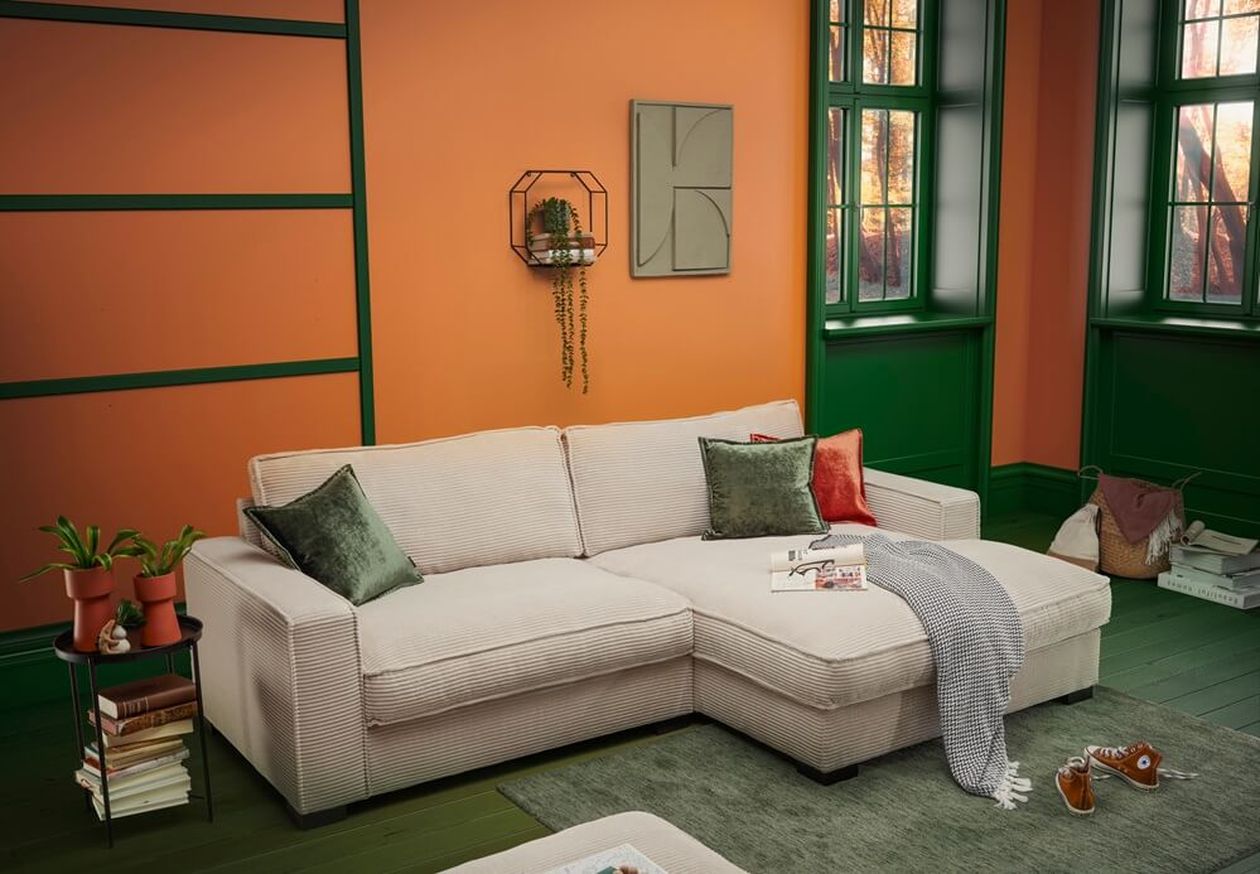 Cremefarbenes Ecksofa aus Cord mit grünen und orangefarbenen Kissen in einem Wohnzimmer mit orangefarbenen Wänden und grünen Fensterrahmen.