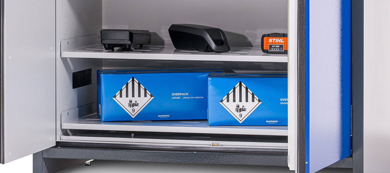 Nyitott szekrény két polccal, amelyeken kék lítium-ion akkumulátordobozok és fekete elektronikus eszközök találhatók. A szekrény ajtaja részben nyitva van.