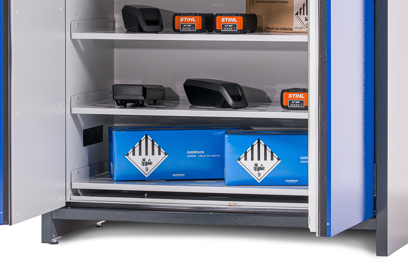 Nyitott szekrény két polccal, amelyeken kék lítium-ion akkumulátordobozok és fekete elektronikus eszközök találhatók. A szekrény ajtaja részben nyitva van.
