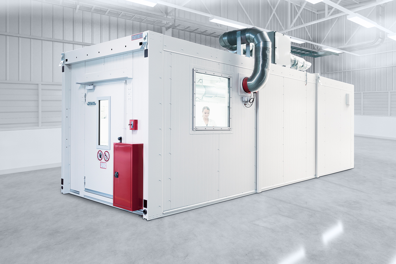 Egy fehér teszthelyiség ajtóval, ablakkal és piros díszítéssel egy nagy raktárban. Felsővezetékek csatlakoznak az egységhez.