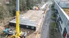 Luchtfoto van een bouwplaats met een grote gele kraan, grijze modulaire gebouwen en nabijgelegen wegen, omgeven door bomen en geparkeerde auto's.