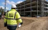 Een bouwvakker in een gele jas en met een veiligheidshelm inspecteert op een zonnige dag een bouwplaats van een gebouw met meerdere verdiepingen, terwijl hij bouwtekeningen vasthoudt.