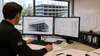 Iemand die aan een bureau werkt met twee beeldschermen waarop bouwtekeningen en een 3D-model te zien zijn, omringd door architecturale plannen en een beker.