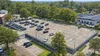 Luchtfoto van een grote parkeerplaats met diverse geparkeerde auto's, omringd door bomen en gebouwen op een zonnige dag.