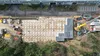 Luchtfoto van een bouwplaats met de contouren van een fundering, bouwvoertuigen en materieel, omgeven door groen en een weg.