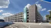 Moderne parkeergarage met meerdere verdiepingen en een gevel van baksteen en glas, onder een halfbewolkte hemel. Een gebogen weg leidt naar de in- en uitrit.
