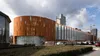 Modern parkeergebouw met een gebogen, roestkleurige gevel en hoge glazen ramen; bewolkte lucht met verspreide wolken en een stadsgezicht van Tilburg op de achtergrond.