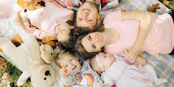 Familie liegt lachend mit Kindern und Teddybär auf einer Picknickdecke im Herbstlaub