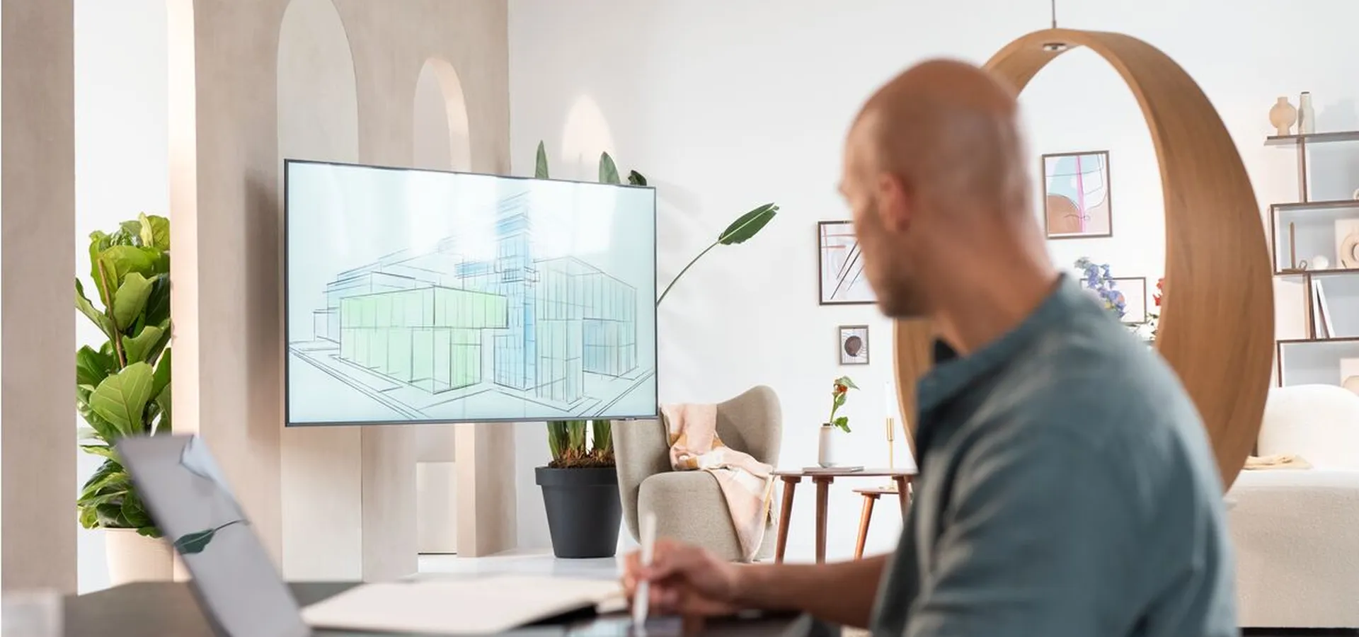 A person working at a desk with a laptop and stylus, viewing architectural designs on a wall-mounted screen in a modern, well-lit room.