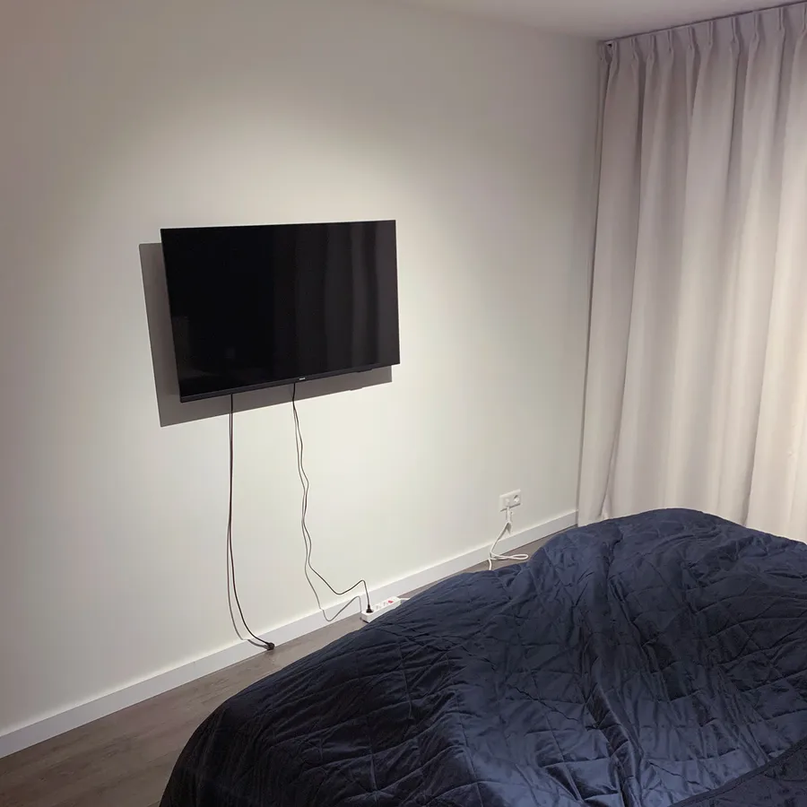 Wall-mounted TV with visible cables above a bed covered in a dark quilt, in a room with light curtains and wooden flooring.