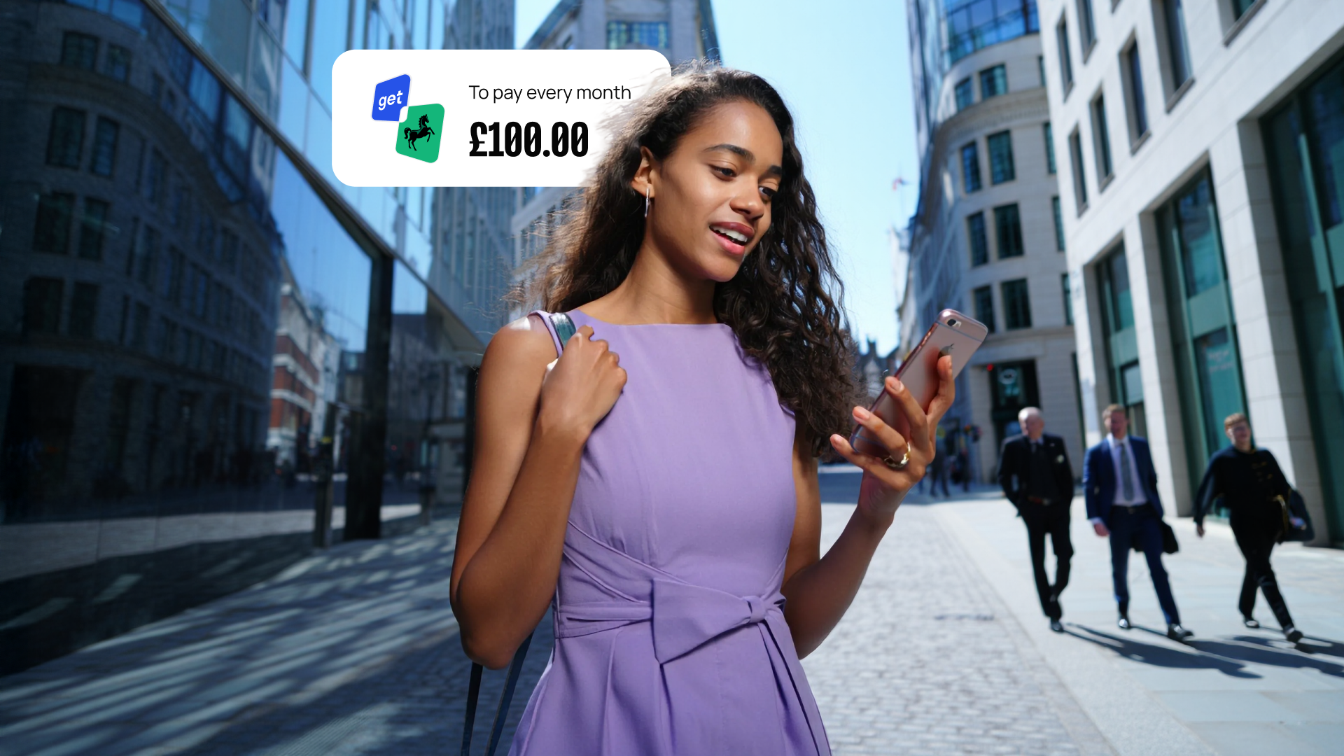 A woman in a purple dress looks at her phone while walking in a city. A notification shows a monthly payment of £108.00.