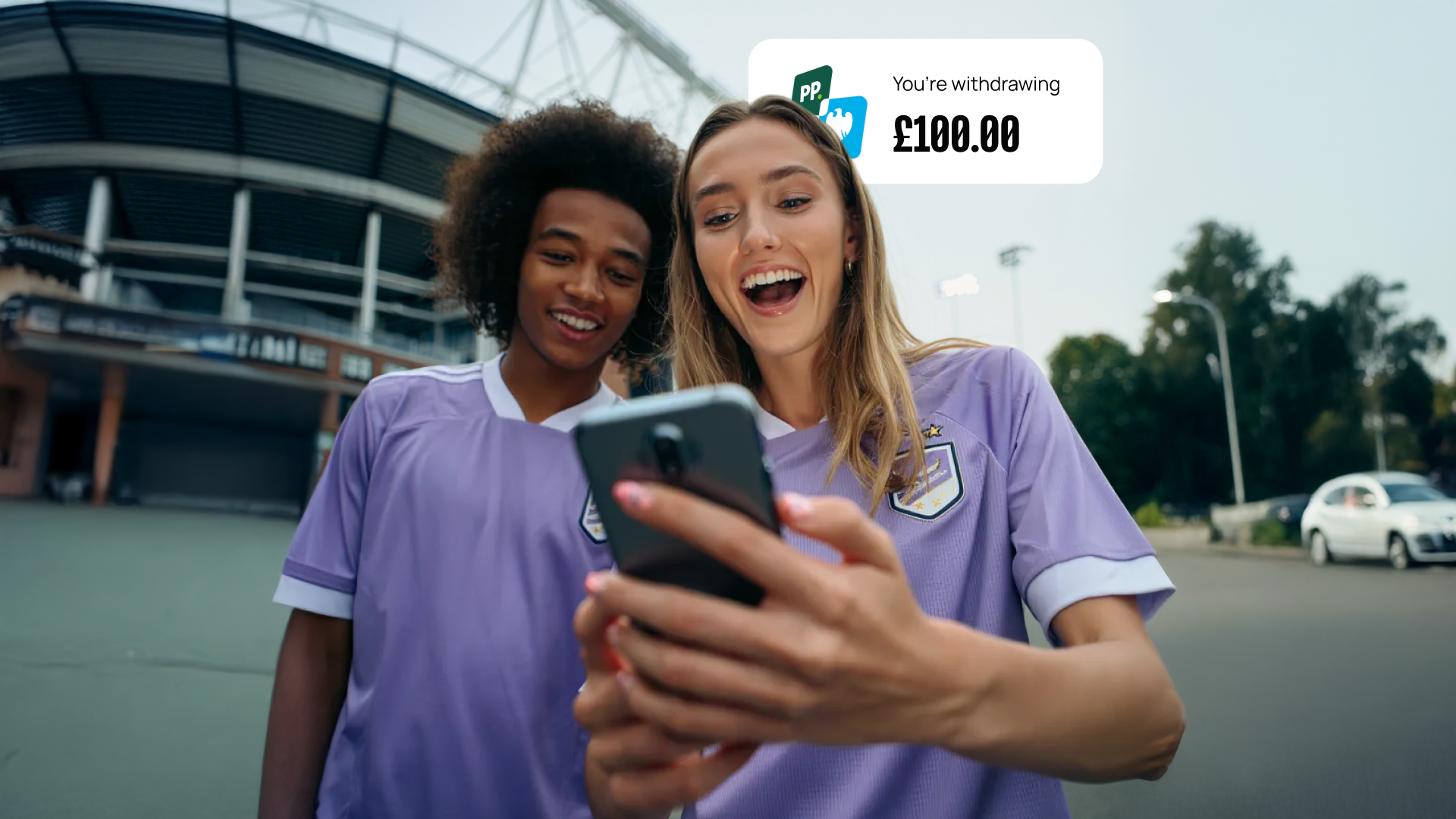 Two people in purple jerseys excitedly looking at a smartphone. A digital notification shows a £100.00 withdrawal. Stadium in the background.