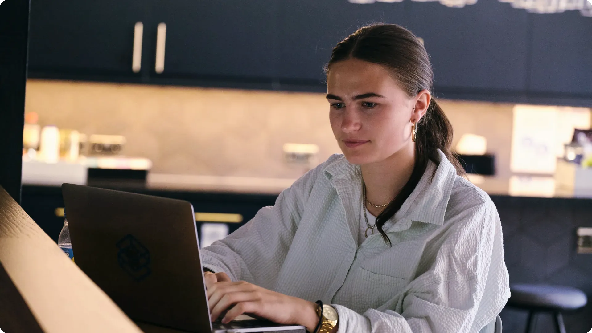 A person with long hair tied back, wearing a white shirt, working intently on a laptop in a dimly lit room.