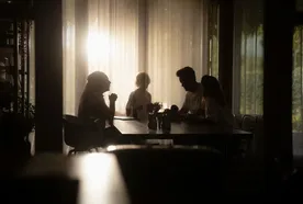 A family sits together around a dining table in a dimly lit room, silhouetted against bright sunlight filtering through sheer curtains.