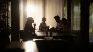 A family sits together around a dining table in a dimly lit room, silhouetted against bright sunlight filtering through sheer curtains.