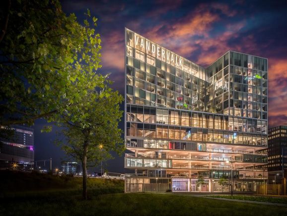 Modern glass building with "VANDERVALK" sign, illuminated at dusk. Tree in foreground, cityscape in background under a colorful sky.