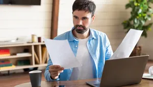 A man in a blue shirt reviews documents at a wooden table with a laptop, smartphone, and coffee cup, in a cozy, well-lit room.