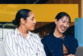 Two women sitting together on a couch, smiling and looking at a laptop screen. One woman wears a striped shirt and has her hair in a bun, while the other wears a dark jacket and has straight hair with bangs. They appear to be having an enjoyable, collaborative conversation in a modern office setting, with colorful graphic overlays in the background.