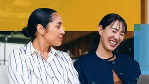 Two women sitting together on a couch, smiling and looking at a laptop screen. One woman wears a striped shirt and has her hair in a bun, while the other wears a dark jacket and has straight hair with bangs. They appear to be having an enjoyable, collaborative conversation in a modern office setting, with colorful graphic overlays in the background.