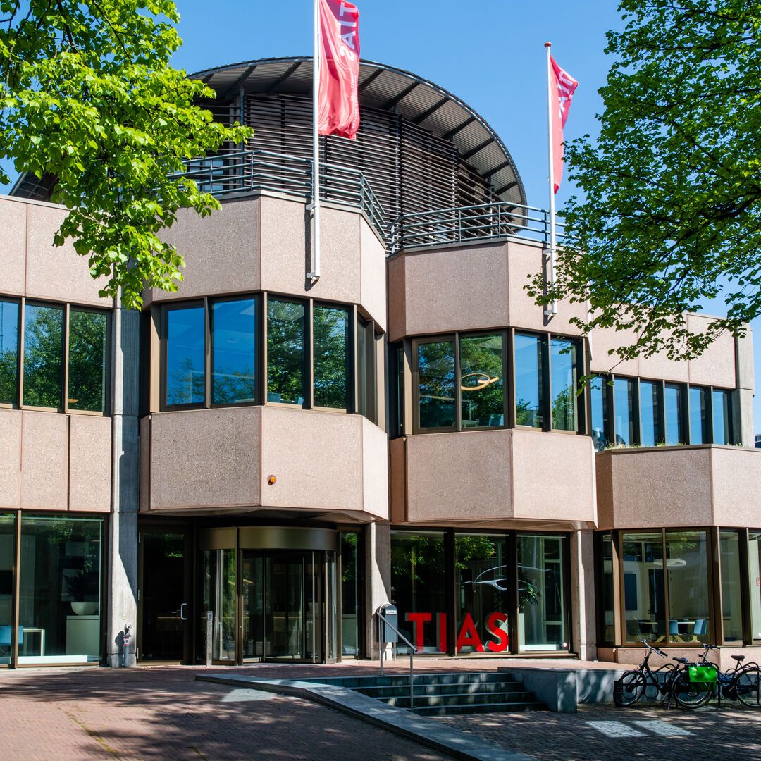 Modern building with glass and beige panels, three red flags on the roof, and red "TIAS" letters at the entrance, surrounded by trees.