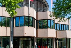 Modern building with glass and beige panels, three red flags on the roof, and red "TIAS" letters at the entrance, surrounded by trees.