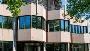 Modern building with glass and beige panels, three red flags on the roof, and red "TIAS" letters at the entrance, surrounded by trees.