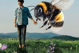 A man in a denim jacket looks at a giant bee hovering beside him in a flowering field, with mountains in the background.