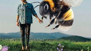 A man in a denim jacket looks at a giant bee hovering beside him in a flowering field, with mountains in the background.