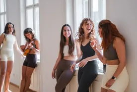 Five women in activewear chat and play guitar near large windows, enjoying a relaxed and cheerful moment in a bright room.