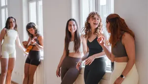 Five women in activewear chat and play guitar near large windows, enjoying a relaxed and cheerful moment in a bright room.
