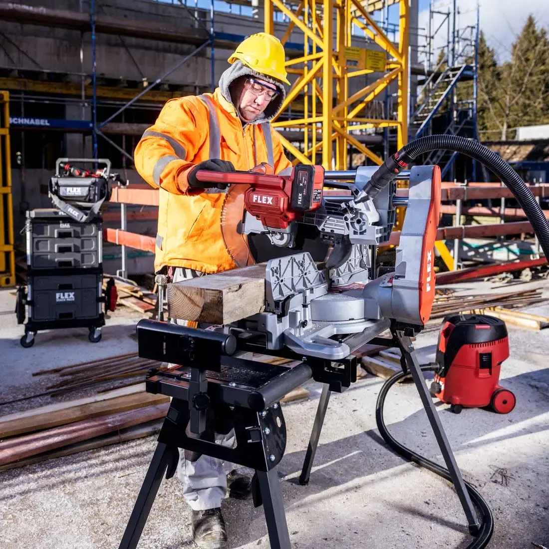 A construction worker wearing an orange safety jacket and yellow hard hat operates a FLEX power saw on a job site. He is cutting a piece of timber using a mounted miter saw, with additional FLEX toolboxes and equipment visible in the background amid scaffolding and building materials.