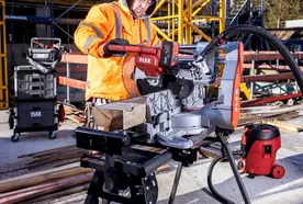 A construction worker wearing an orange safety jacket and yellow hard hat operates a FLEX power saw on a job site. He is cutting a piece of timber using a mounted miter saw, with additional FLEX toolboxes and equipment visible in the background amid scaffolding and building materials.