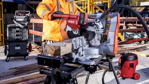 A construction worker wearing an orange safety jacket and yellow hard hat operates a FLEX power saw on a job site. He is cutting a piece of timber using a mounted miter saw, with additional FLEX toolboxes and equipment visible in the background amid scaffolding and building materials.