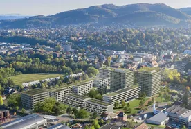 Aerial view of a cityscape with modern buildings, green spaces, and surrounding hills under a clear blue sky.
