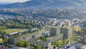 Aerial view of a cityscape with modern buildings, green spaces, and surrounding hills under a clear blue sky.