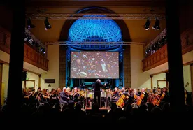 Orchestra performing in a grand hall with a large screen projection of colorful objects above the stage, illuminated by blue lighting.