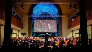 Orchestra performing in a grand hall with a large screen projection of colorful objects above the stage, illuminated by blue lighting.