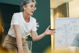 Person with short hair presenting at a meeting, gesturing animatedly, with a flowchart on a whiteboard in the background.