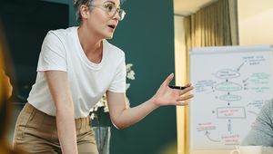 Person with short hair presenting at a meeting, gesturing animatedly, with a flowchart on a whiteboard in the background.