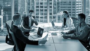 Five professionals in a modern office meeting room with laptops and documents, cityscape visible through large windows in the background.