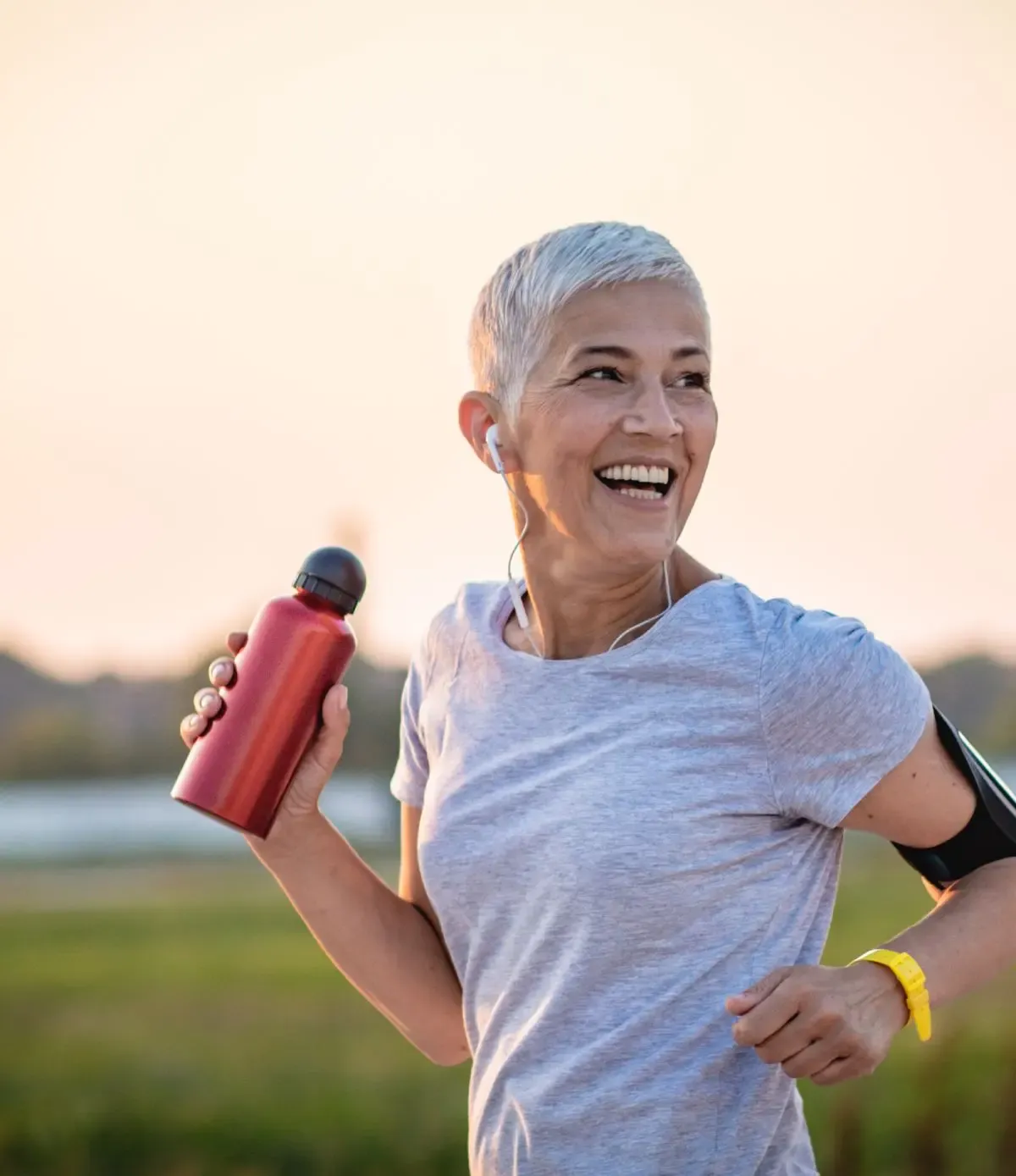 Smiling woman with short gray hair runs outdoors, holding a red water bottle. She wears earbuds, a gray shirt, and an armband at sunset.