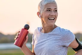 Smiling woman with short gray hair runs outdoors, holding a red water bottle. She wears earbuds, a gray shirt, and an armband at sunset.