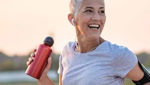Smiling woman with short gray hair runs outdoors, holding a red water bottle. She wears earbuds, a gray shirt, and an armband at sunset.