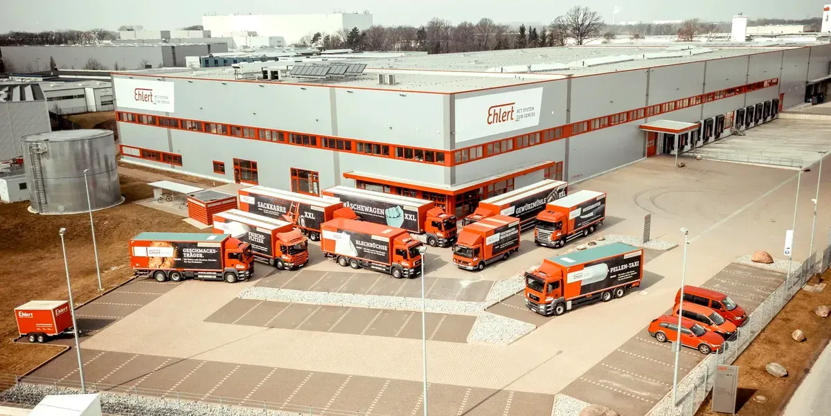 Aerial view of a logistics warehouse with multiple red and white trucks and vans parked in front, and a few red cars nearby.