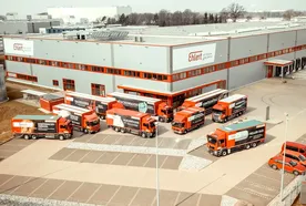 Aerial view of a logistics warehouse with multiple red and white trucks and vans parked in front, and a few red cars nearby.