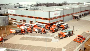 Aerial view of a logistics warehouse with multiple red and white trucks and vans parked in front, and a few red cars nearby.