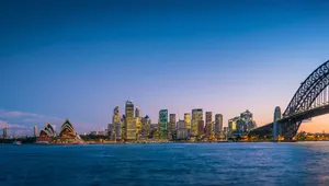 Skyline of Sydney at dusk, featuring the Opera House, lit skyscrapers, and the Harbour Bridge over calm water.