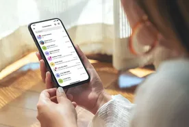 Person holding a smartphone displaying a list of messages in a messaging app, sitting in a sunlit room.