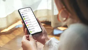 Person holding a smartphone displaying a list of messages in a messaging app, sitting in a sunlit room.