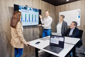 A person presents a bar chart on a screen to three colleagues in a meeting room with laptops and a whiteboard.